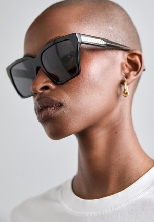 Sonnenbrille mit einem dicken schwarzen Rahmen, dunklen Gläsern und goldfarbenen Akzenten an den Bügeln, die ein strukturiertes Muster aufweisen. Glatte Haut und ein einfaches weißes Shirt sind sichtbar.