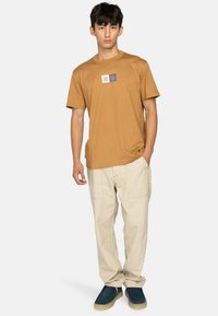 Camiseta de algodón marrón con un parche de logo cuadrado en blanco y azul. Combinada con pantalones crema claros y zapatillas azules sin cordones. Diseño simple y casual.