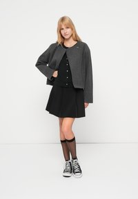 Γκρι κοντό σακάκι με φαρδιά μανίκια, μαύρη μπλούζα με κουμπιά, μαύρη φούστα με φουσκωτό σχέδιο, μαύρα καλσόν και μαύρα αθλητικά παπούτσια.