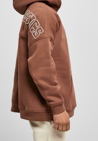 Sudadera de algodón marrón con un bolsillo frontal y el texto en blanco "CLASSICS" en la manga izquierda. Presenta un ajuste regular y puños de canalé.
