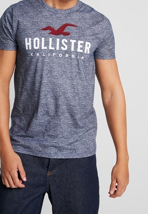 Mężczyzna w niebieskim melanżowym t-shircie Hollister California z czerwonym logo ptaka i ciemnoniebieskich dżinsach, stojący na białym tle.