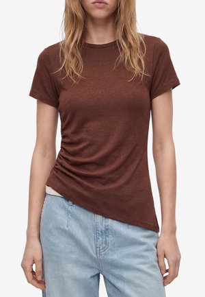Donna che indossa una t-shirt marrone arricciata a maniche corte e jeans azzurri con lunghi capelli biondi su sfondo bianco.