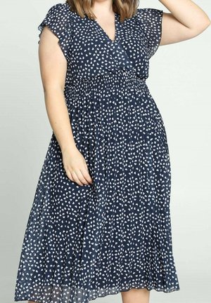 Kobieta w granatowej sukience midi z białym wzorem serduszek, z falbankami na rękawach i marszczonym pasem, stojąca na jasnym tle.