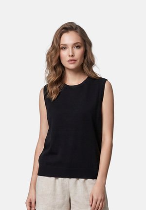 Junge Frau mit welligem Haar, die ein ärmelloses schwarzes Top und hellbeige Shorts trägt und vor einem schlichten hellen Hintergrund steht.