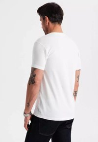 T-shirt bianco in cotone con maniche corte e collo rotondo, mostrato da dietro. Presenta tatuaggi visibili sul braccio.