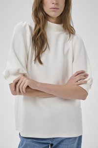 Ženska z dolgimi rjavimi lasmi, oblečena v belo bluzo z napihanimi rokavi in modre kavbojke, z rokami prekrižanimi v pasu, nevtralno sivo ozadje.