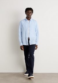 Lichtblauwe linnen overhemd met knopen, donkere marineblauwe broek en zwarte sneakers, met een slim fit en casual stijl, staand tegen een neutrale achtergrond.