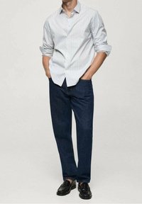 Camicia bianca con righe verticali blu e rosse, abbinata a pantaloni in denim blu scuro e mocassini neri. Vestibilità casual con maniche arrotolate.