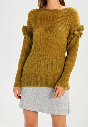 Mujer con un suéter acanalado color mostaza con detalles de volantes en las mangas y una falda gris claro hasta la rodilla.