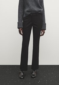 Pantalon évasé noir à texture lisse, avec une fermeture classique à boutons et des poches latérales, associé à un pull gris côtelé.