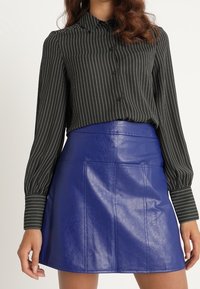 Schwarz gestreiftes Hemd mit langen Ärmeln, kombiniert mit einem hochgeschnittenen, glänzenden blauen Leder-Minirock. Glatte Textur und taillierte Passform.