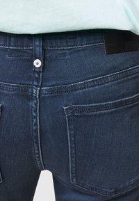 Mörkblå denimjeans med en mjuk textur, med en silverfärgad knäppning, två bakfickor och en distinkt svart läderlogotyp.