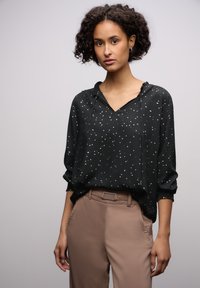 Musta paita kultaisella pilkkukuviolla, v-aukolla ja nyöri hupulla. Rentohenkinen malli, jossa on käärityt hihat, yhdistetty vaaleanruskeisiin housuihin.