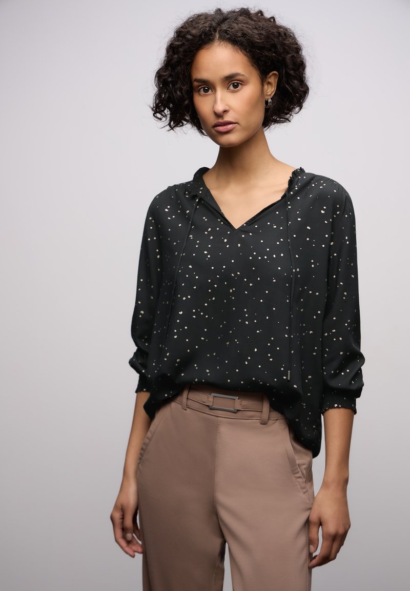 Musta paita kultaisella pilkkukuviolla, v-aukolla ja nyöri hupulla. Rentohenkinen malli, jossa on käärityt hihat, yhdistetty vaaleanruskeisiin housuihin.