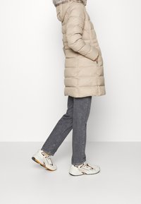 Beige pufferjack met capuchon en bontafwerking, met een gestikte uitstraling. Gecombineerd met grijze jeans en witte sneakers met accenten.