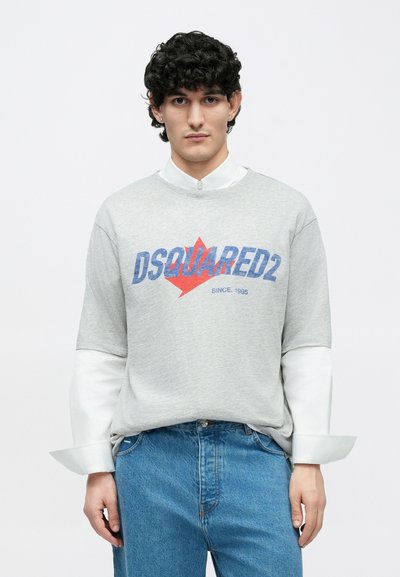 Grauer Sweatshirt mit rotem und blauem "DSQUARED2"-Logo und Ahornblatt-Grafik. Weiße innere Bündchen sichtbar. Kombiniert mit blauen Denim-Jeans.