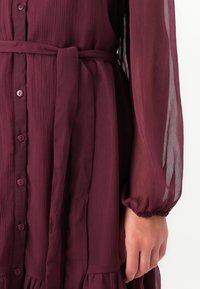 Close-up of a deep burgundy button-up dress with a tied waist belt and sheer long sleeves on a person's arm and hand.
