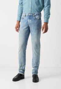 Vaqueros de denim azul claro con un corte ajustado, que presentan un ligero desgaste y diseño de cinco bolsillos. Combinados con mocasines de cuero negros.