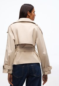 Frau mit langen, dunklen Haaren, die einen beigen Trenchcoat mit Gürtel und dunkelblaue Jeans trägt, von hinten vor weißem Hintergrund gesehen.