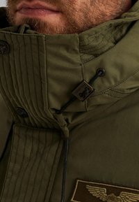 Maslinasto zelena jakna sa patentnim zatvaračem i visokim ovratnikom, s prešanim teksturom, tamnim šavovima, oznakom brenda i podesivim vezanjem.