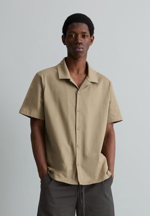 Młody mężczyzna stoi z rękami w kieszeniach, ubrany w beżową koszulę na krótki rękaw z guzikami oraz ciemnoszare spodnie, na jednolitym tle.