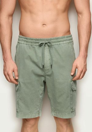 Mężczyzna w jasnych zielonych szortach cargo z elastycznym paskiem i sznurkiem, stojący z rękami swobodnie opuszczonymi wzdłuż ciała.