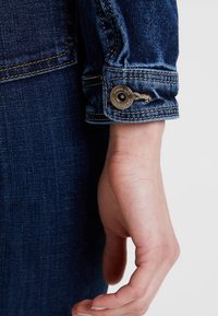 Manžeta tmavě modré džínové bundy s kontrastním žlutým šitím a kovovým patentním knoflíkem s vyrytým logem. Hladká textura.