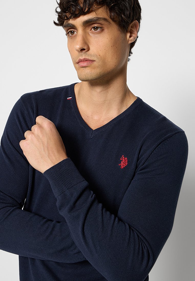 Marineblaues V-Ausschnitt-Pullover aus Strickstoff, mit einem roten gestickten Logo auf der Brust und gerippten Bündchen für Textur und Passform.