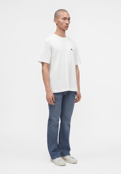 Homme debout avec une expression neutre, portant un t-shirt blanc à manches courtes, un jean bleu et des baskets blanches sur un fond blanc uni.