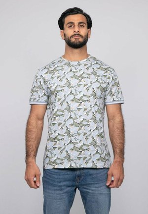 Homme barbu portant un t-shirt blanc à manches courtes avec un motif de feuilles vertes et un jean bleu, debout devant un fond clair uni.