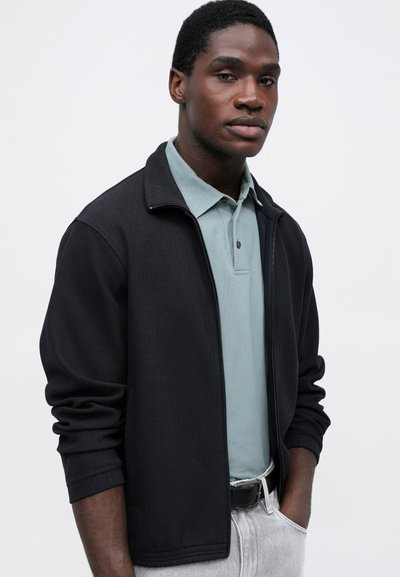 Čierna zipsová bunda s herringbonovým vzorom, nosená cez svetlomodrý polo tričko. Jednoduchý dizajn, dlhé rukávy a voľný strih.