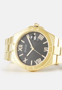 Montre dorée avec un cadran facetté, un cadran gris foncé, des chiffres romains et un affichage de la date. Bracelet en métal doré attaché avec des maillons.