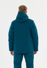 Mann mit kurzen blonden Haaren, der eine türkisfarbene Kapuzenjacke und passende Hosen trägt, steht von hinten vor einem schlichten weißen Hintergrund.