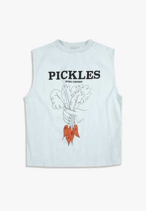 Jeune fille debout portant un débardeur avec le texte "PICKLES" et une illustration de légumes, un short fleuri, des chaussettes blanches et des sandales noires.