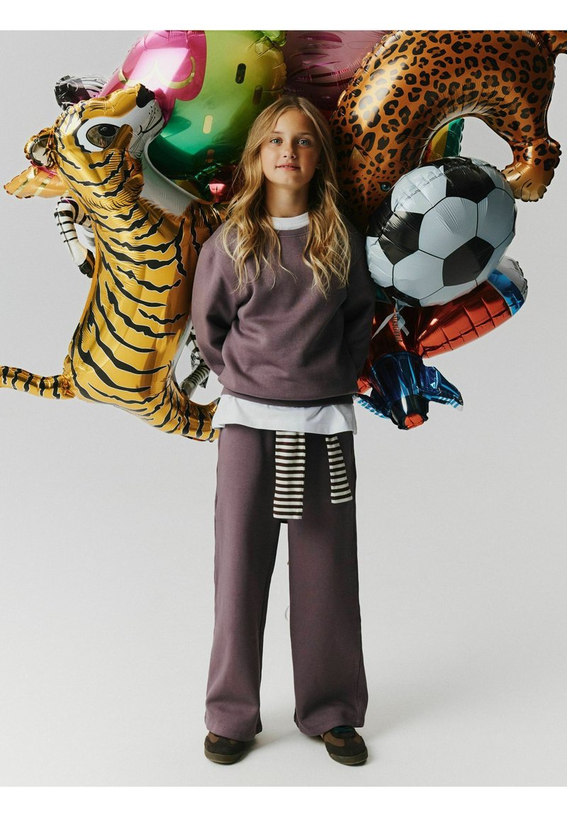 Fille aux longs cheveux blonds portant un pantalon de survêtement gris et un sweat-shirt, debout avec des ballons colorés en forme d'animaux et de ballon de football derrière elle.