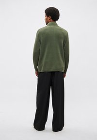 Πουλόβερ σε ελαιώδες πράσινο με ψηλό γιακά, μακριά μανίκια και αντίθετες ραφές, συνδυασμένο με μαύρο παντελόνι φαρδύ στο τελείωμα.