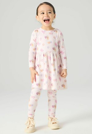 Jauna mergina juokiasi, dėvi ilgomis rankovėmis suknelę ir leginsus su pasteliniais ledų ragelių raštais bei smėlio spalvos sportbačius, stovinti ant paprasto fono.