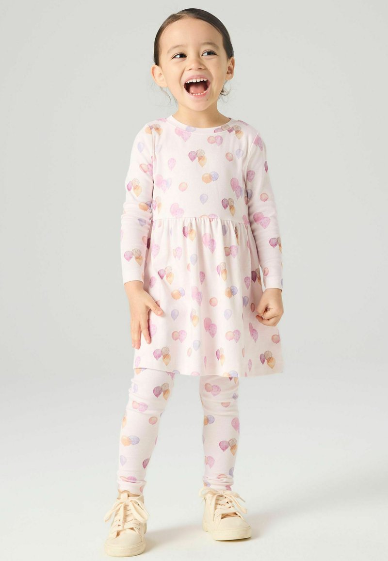 Ung jente som ler, iført en langermet kjole og leggings med pastellfargede iskremkjeglemønstre og beige joggesko, stående på en ensfarget bakgrunn.