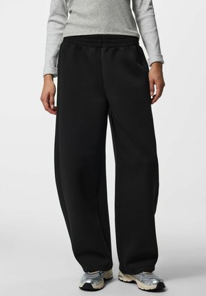 Personne portant un pantalon noir ample, un haut gris à manches longues et des chaussures de course argentées, debout devant un fond uni.