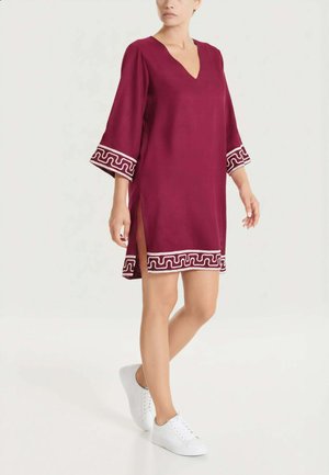 Donna con indosso un vestito bordeaux a scollo a V con rifiniture geometriche bianche su polsini e orlo, abbinato a sneakers bianchi, in piedi su un pavimento bianco.