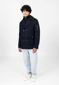 Granatowy wełniany podwójnie zapięty płaszcz peacoat z dużymi guzikami, bocznymi kieszeniami i kieszenią na zamek na rękawie, w zestawieniu z jasnoniebieskimi dżinsami i białymi trampkami.