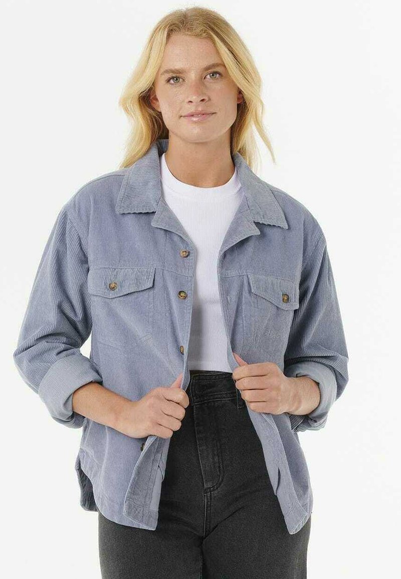 Giacca in corduroy azzurra chiara con chiusura a bottoni, due tasche sul petto e maniche arrotolate. Indossata sopra una camicia bianca e pantaloni neri.