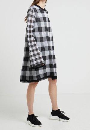 Kvinde iført en sort-hvid ternet langærmet kjole kombineret med sorte sokkesneakers med hvide såler mod en ensfarvet baggrund.