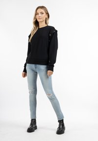 Zwarte sweatshirt met schouderaccenten en knoopdetails, in combinatie met lichtblauwe ripped jeans en zwarte enkellaarsjes.
