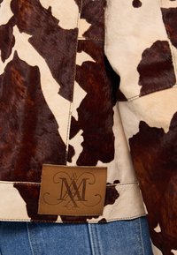 Brun och krämfärgad kohudarjacka med en texturerad yta, utrustad med en läderetikett med präglade initialer. Jeans synliga nedanför.