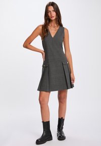 Harmaan ruudullinen mekko V-pääntiellä, hihaton muotoilu, laskostettu hame sekä kaksinkertainen etutasku napitusyksityiskohdilla, yhdistetty mustiin nilkkasaappaisiin.