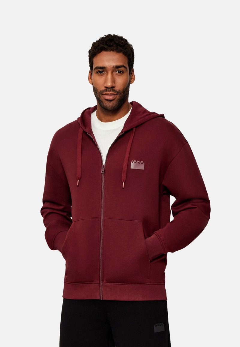 Sudadera con cremallera en color burdeos, hecha de tejido suave, con un bolsillo frontal, capucha ajustable con cordón y acento de logo en el pecho.