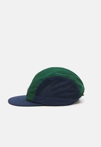 Gorra verde y azul marino con paneles laterales de malla y visera plana, vista de perfil sobre un fondo blanco liso.