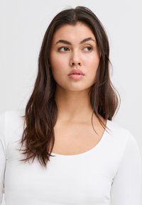 Fehér, hosszú ujjú felső kerek nyakkivágással, sima textúrával, egy hosszú, sötét hajú személy által viselve, világos háttér előtt.