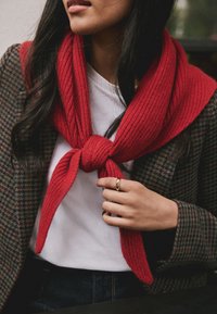 Sciarpa rossa in maglia a costine annodata intorno a una camicia bianca, abbinata a un blazer a fantasia. Mano visibile che indossa un anello d'oro.